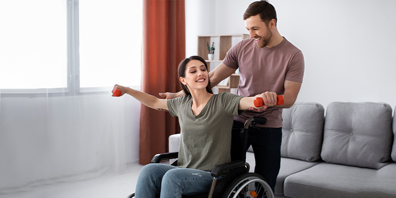 Woman in wheelchair exercising with weights, assisted by a caregiver, promoting daily living skills support for independent living.